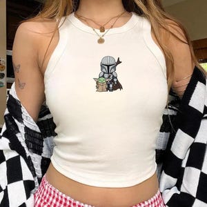 May include: White tank top with a graphic of The Mandalorian and Grogu. The sleeveless top has a high neckline and a fitted silhouette. The graphic is in the center of the top and features the characters in a cartoon style.