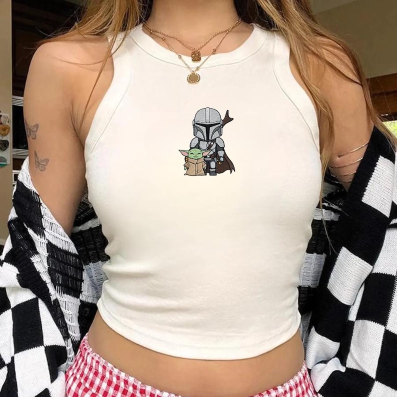 May include: White tank top with a graphic of The Mandalorian and Grogu. The sleeveless top has a high neckline and a fitted silhouette. The graphic is in the center of the top and features the characters in a cartoon style.
