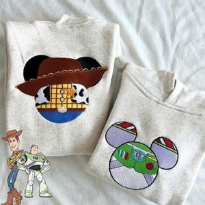 May include: Two light gray hoodies with embroidered cartoon character designs. One features a brown cowboy hat and a sheriff's badge, while the other has a green and purple space ranger design. Both designs are in the shape of Mickey Mouse ears.