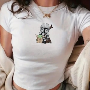 May include: White short-sleeved t-shirt featuring a cartoon illustration of The Mandalorian and Grogu. The graphic shows the characters in full armor and robes. The shirt is made of a soft, lightweight material.