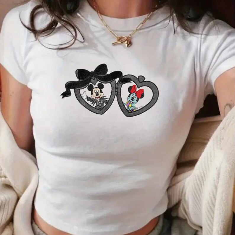May include: A white short-sleeved t-shirt featuring a graphic of two heart-shaped lockets connected by a black bow. The left locket contains a black and white character illustration, while the right locket displays a colorful character with a red bow.