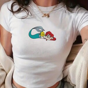 May include: White t-shirt featuring a cartoon illustration of a mermaid with red hair and a teal tail, accompanied by a yellow fish. The graphic is centered on the front of the short-sleeved shirt.