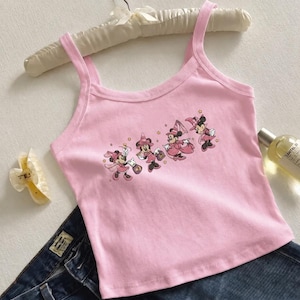 May include: A light pink sleeveless tank top featuring a graphic print of four Minnie Mouse characters dressed in pink wizard hats and robes, accented with small yellow stars. The top has a scoop neckline and wide shoulder straps.