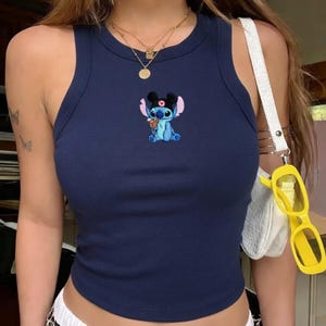 Puede incluir: Camiseta sin mangas de canal&eacute; azul marino con un dise&ntilde;o bordado del personaje Stitch. La parte superior sin mangas tiene un cuello redondo y una silueta ajustada. Un bolso blanco con gafas de sol amarillas cuelga a un lado.
