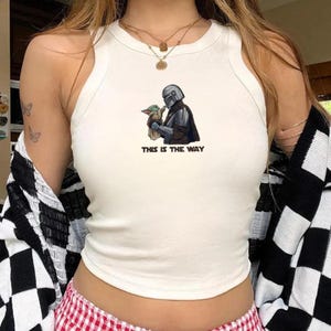 May include: White tank top featuring a graphic of a Mandalorian character holding Grogu, with the text "THIS IS THE WAY" in black. The image is centered on the front of the top.