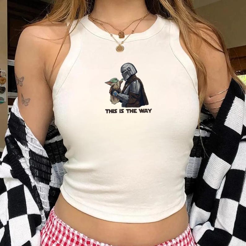 May include: White tank top featuring a graphic of a Mandalorian character holding Grogu, with the text "THIS IS THE WAY" in black. The image is centered on the front of the top.
