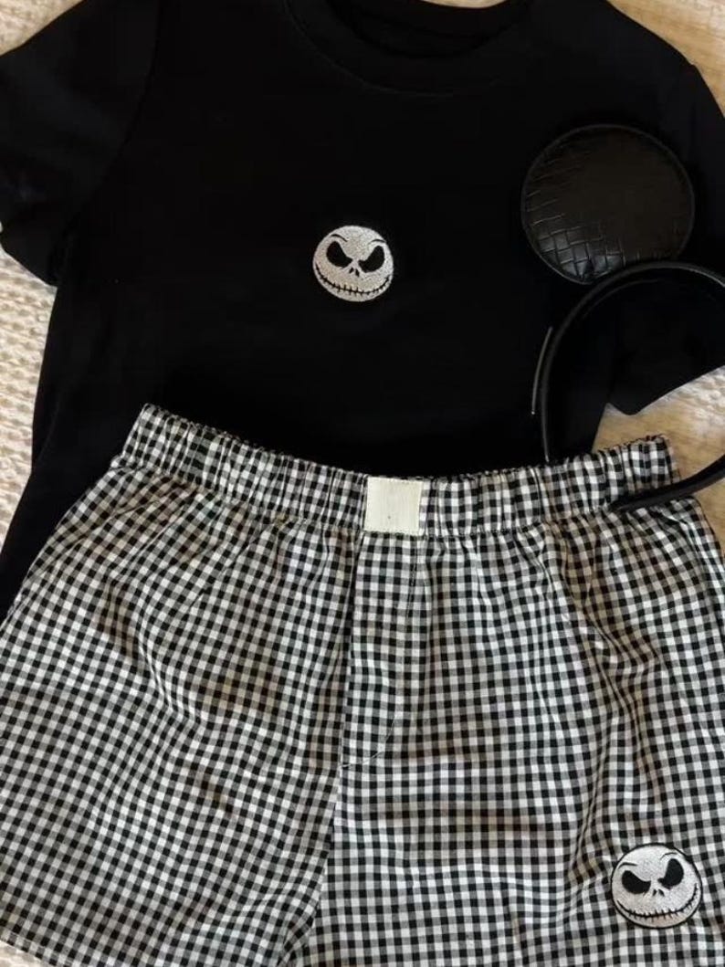 May include: A black t-shirt with a Jack Skellington face patch, black and white checked boxer shorts, and a black mouse ear headband. The shorts also feature a Jack Skellington face patch. This is likely a themed sleepwear set.