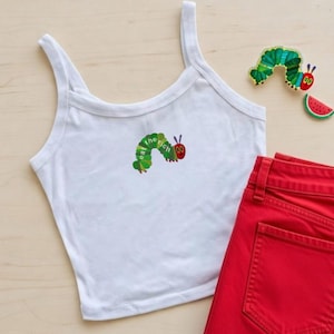 Könnte beinhalten: Weißes Tanktop mit einem grünen und roten Raupen-Grafik, auf dem "eat the rich" steht. Rote Jeansshorts liegen neben dem Tanktop. Ein Raupen- und Wassermelonen-Patch sind ebenfalls im Bild.