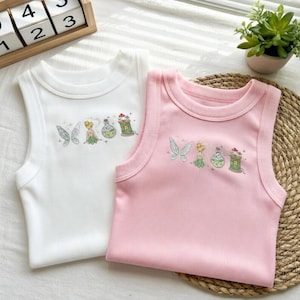 Könnte beinhalten: Zwei ärmellose Shirts, eines weiß und eines rosa, werden gezeigt. Jedes Shirt hat ein gesticktes Design einer Fee, Schmetterlingsflügeln und Zaubertränken. Die Shirts sind aus einem weichen Rippstoff gefertigt.