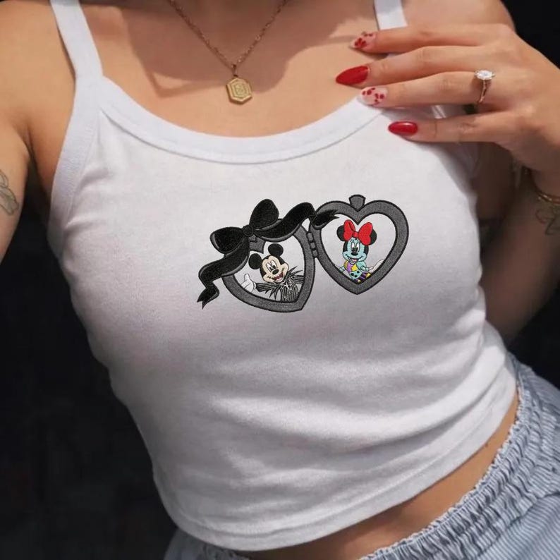 May include: A white tank top featuring a graphic print of two heart-shaped lockets connected by a black bow. The left locket contains an illustration of Mickey Mouse, and the right locket contains an illustration of Minnie Mouse with a red bow.