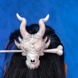May include: A white dragon skull hair clip with a bone-like design. The clip is attached to a wooden stick.