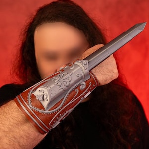 Può includere: Una persona che indossa una camicia nera e un braccialetto da polso in pelle marrone con accenti argentati. Il braccialetto ha un design decorativo e un piccolo pezzo di metallo sul lato.