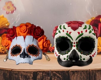 May include: Two decorative sugar skull masks, one light blue and one white, sit on a wooden surface. The light blue skull has orange and red roses. The white skull has a black mustache and is decorated with red and green accents.