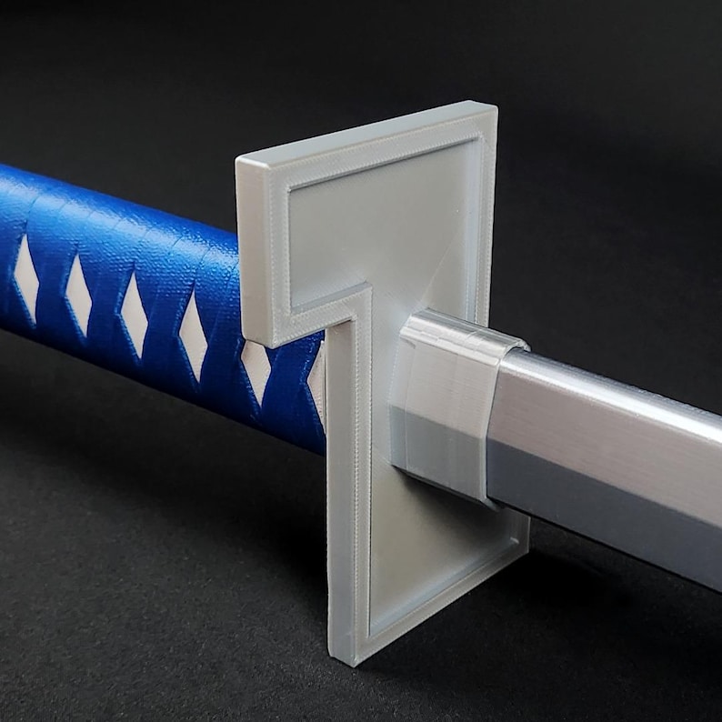 May include: Close-up of a katana sword. The handle is wrapped in blue fabric with white diamond patterns. The guard is a light gray color, and the blade is silver. The sword is set against a dark background.