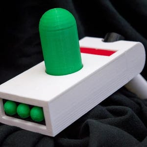 May include: A white plastic toy gun with a green cylindrical projectile and a red stripe. The gun has a row of five green spheres on the bottom.