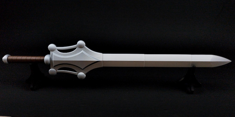 May include: A sword with a long white blade and a pointed tip. The hilt features a white crossguard with four circular accents and a brown, textured grip. The sword is positioned horizontally and is supported by two small black stands.