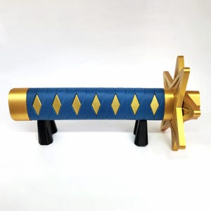 May include: A decorative katana handle with a blue grip featuring gold diamond accents. The hilt and guard are gold, and the handle rests on a black stand. The overall design is a stylized representation of a traditional Japanese sword.