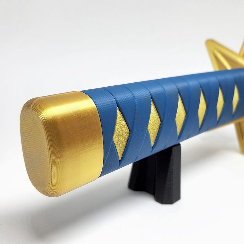 May include: A close-up of a blue and gold katana handle. The handle is wrapped in blue with gold diamond-shaped accents. The end cap is gold. The katana is resting on a black stand.