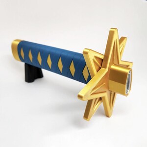 May include: A gold and blue decorative sword. The handle is blue with yellow diamond accents. The guard is a gold star shape. The sword is resting on a black stand.