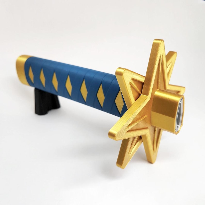 May include: A gold and blue decorative sword. The handle is blue with yellow diamond accents. The guard is a gold star shape. The sword is resting on a black stand.