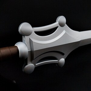 May include: A sword with a silver-colored crossguard featuring four rounded circular accents at the corners and a brown textured handle. The blade extends from the center of the guard, which has a smooth, metallic finish and visible layered lines.