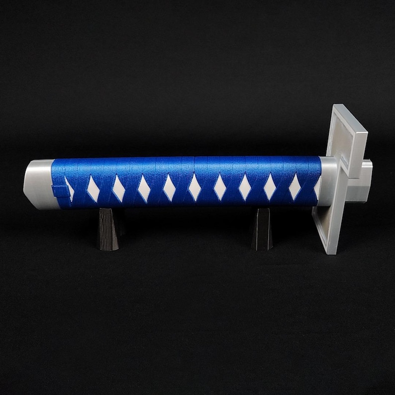May include: A katana handle with a blue grip wrapped with white diamond-shaped accents. The hilt is silver, and the sword rests on a black stand against a black background. A decorative item.