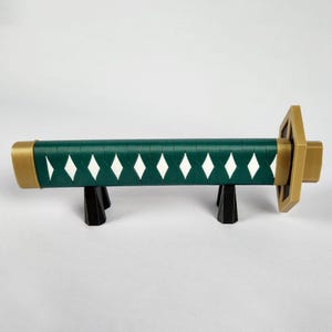 May include: A decorative katana handle with a dark green grip, white diamond accents, and gold-colored accents on the ends and guard. The handle rests on a black stand, showcasing the design and craftsmanship.