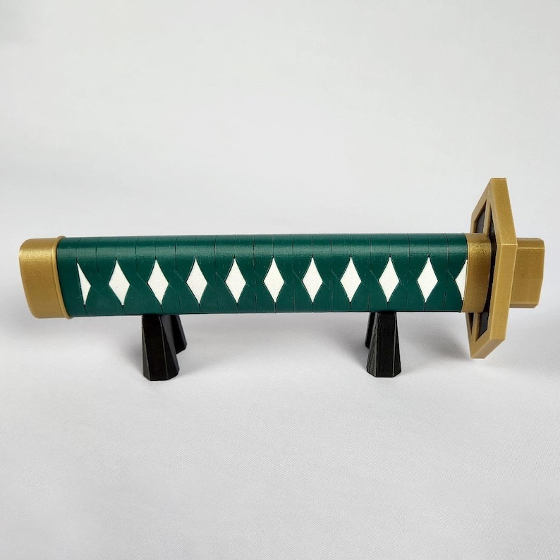 May include: A decorative katana handle with a dark green grip, white diamond accents, and gold-colored accents on the ends and guard. The handle rests on a black stand, showcasing the design and craftsmanship.