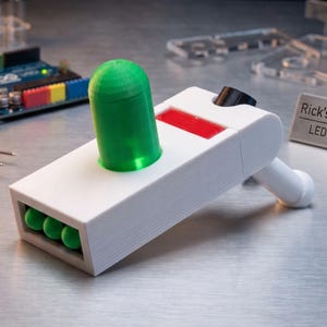 Puede incluir: Una réplica de utilería blanca de la pistola de portal de Rick con una parte superior verde, detalles rojos y esferas verdes. El accesorio tiene el texto "Rick's Portal Prop LED integrated" en un letrero pequeño. Otros componentes electrónicos y herramientas están en el fondo.