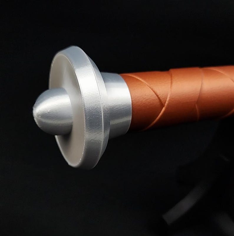 May include: A sword pommel featuring a smooth, silver-colored, rounded cap attached to a cylindrical handle wrapped in a textured, copper-colored material. The handle exhibits a diagonal, crisscross pattern along its length, leading to the pommel.