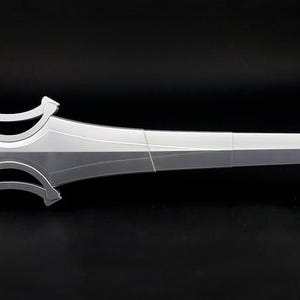 May include: A silver-colored sword with a wide, stylized crossguard and a brown, textured handle. The blade features a central ridge and a pointed tip. The sword is displayed horizontally, supported by two small black stands against a dark background.