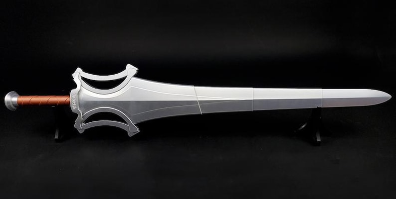 May include: A silver-colored sword with a wide, stylized crossguard and a brown, textured handle. The blade features a central ridge and a pointed tip. The sword is displayed horizontally, supported by two small black stands against a dark background.