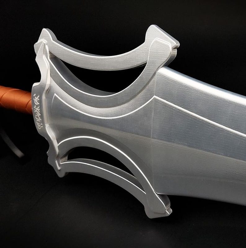 May include: A silver-colored sword with a wide, flat blade and a complex, open-work guard featuring curved geometric cutouts. The handle is wrapped in a smooth, brown material, and the guard displays engraved, stylized markings on its side.
