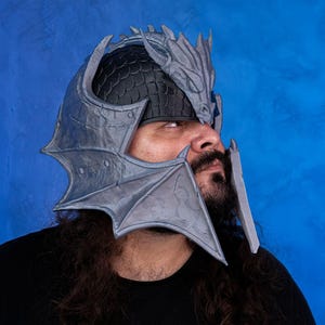 May include: A grey 3D printed dragon helmet with a textured surface. The helmet has a dragon head on top and wings on the sides.