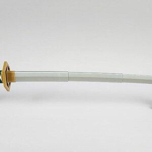 May include: A katana sword with a green handle featuring white diamond-shaped cutouts. The sword has a gold-colored handguard and a silver blade. It is displayed on two black stands against a white background.