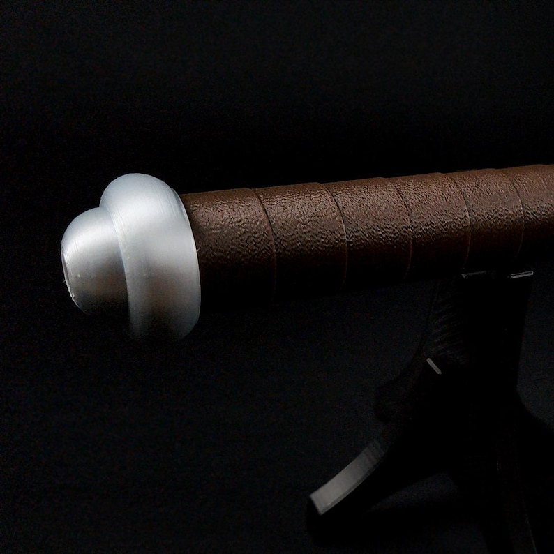 May include: A sword handle featuring a brown textured grip wrapped in a spiral pattern and a smooth, rounded silver-colored pommel at the end. The cylindrical handle is positioned horizontally, showcasing the contrast between the metallic end and the grip.