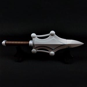 May include: A silver-colored prop weapon featuring a pointed blade with a wide, decorative guard adorned with four spherical accents. The handle is wrapped in a brown textured material, and the pommel is a smooth, rounded silver-colored cap.