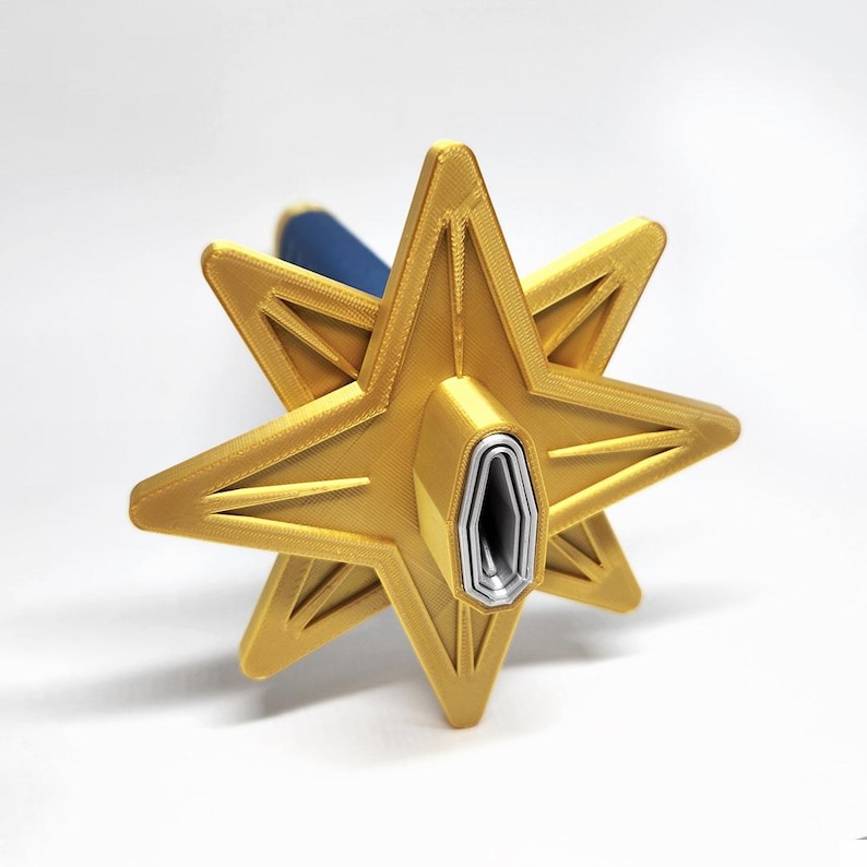 May include: A gold-colored, eight-pointed star-shaped object with a blue handle. The star has a detailed, geometric design with a hollow center. The object appears to be 3D-printed and sits against a white background.