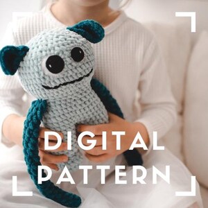 May include: A crocheted plush toy in shades of light blue and teal, held by a person. The toy has large black eyes, a stitched smile, and teal accents on its ears and limbs. The text "DIGITAL PATTERN" is overlaid on the image.