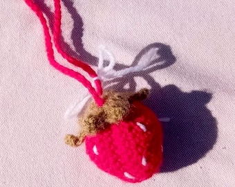 May include: A handmade, crocheted strawberry charm. The bright red fruit has white seed details and a brown stem. A pink and white string is attached for hanging. The charm is set against a pink fabric background.