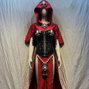May include: A red hooded cloak and black corset costume featuring silver fleur-de-lis emblems, a central skull pendant, a pearl-like beaded chain, and a long skirt with silver grommets and white skull-and-cross symbols printed on the lower hem.