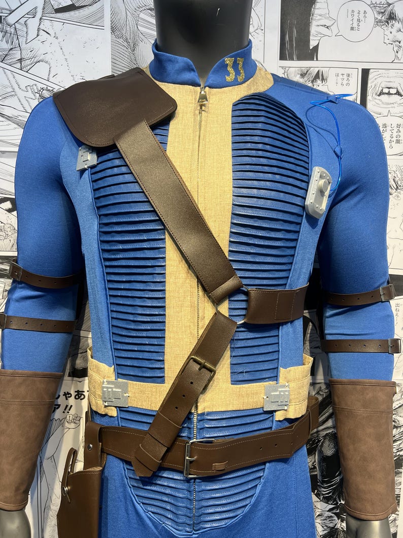 May include: A blue and tan jumpsuit with brown leather straps and a belt. The jumpsuit has a zipper in the front and a patch on the left shoulder. The jumpsuit is worn by a mannequin.