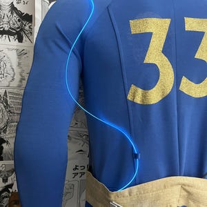 May include: A blue jumpsuit with a gold number 33 on the back. A blue glowing wire is attached to the jumpsuit and wraps around the wearer's torso. The jumpsuit has a tan fabric belt with a silver buckle.