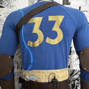 May include: A blue jumpsuit with a brown leather strap and a gold glitter number 33 on the back. The jumpsuit has a brown belt with a silver buckle and a black pouch attached to the belt.