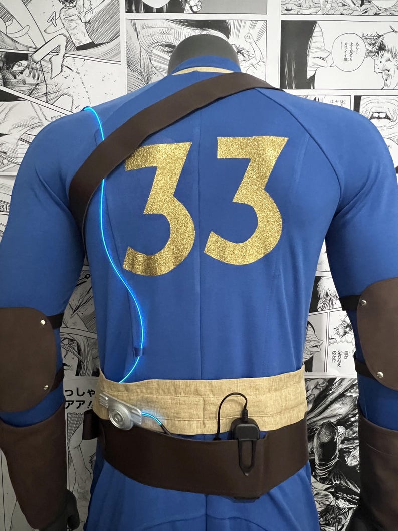 May include: A blue jumpsuit with a brown leather strap and a gold glitter number 33 on the back. The jumpsuit has a brown belt with a silver buckle and a black pouch attached to the belt.