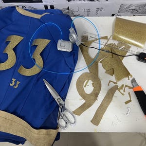 May include: A blue jersey with the number 33 in gold glitter, a pair of scissors, a roll of gold glitter fabric, and a small electronic device with wires.