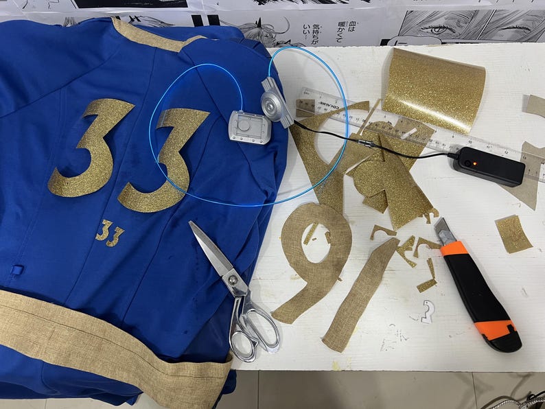 May include: A blue jersey with the number 33 in gold glitter, a pair of scissors, a roll of gold glitter fabric, and a small electronic device with wires.
