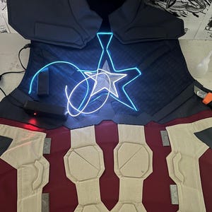May include: A blue and red fabric costume vest with a white star and blue LED lights. The vest has a quilted design and is made of a thick fabric.