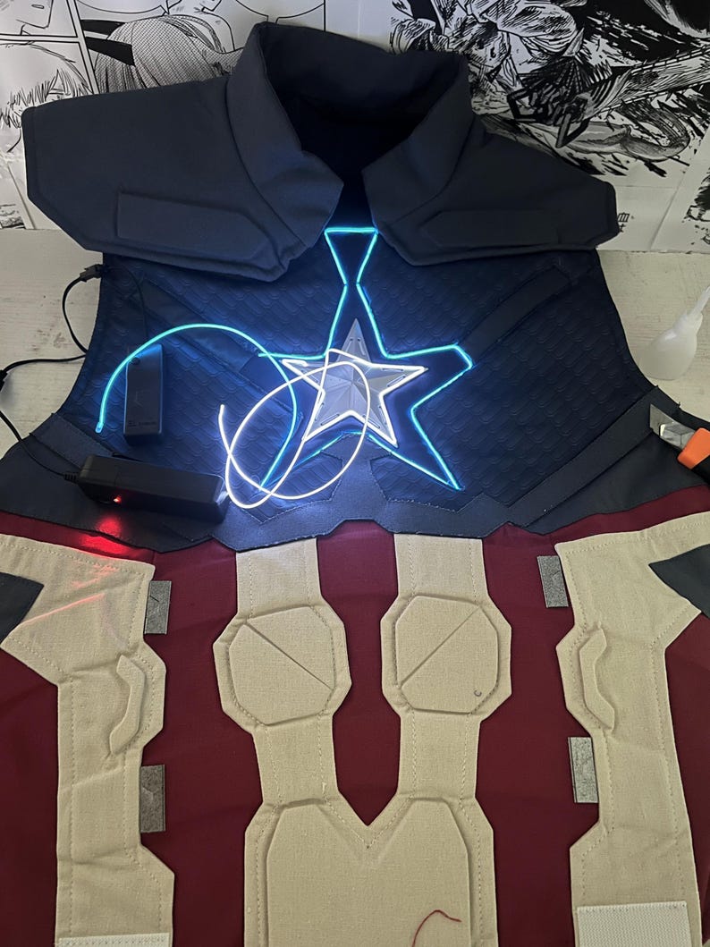 May include: A blue and red fabric costume vest with a white star and blue LED lights. The vest has a quilted design and is made of a thick fabric.