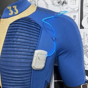 May include: A blue jumpsuit with a tan collar and a silver badge on the chest. The jumpsuit has a brown belt and a blue light cord running from the shoulder to the badge. The number "33" is embroidered on the collar in gold thread.